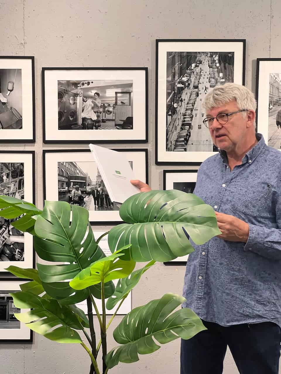 Ein älterer Mann in einem blauen Hemd steht in einer Kunstgalerie und betrachtet verschiedene schwarz-weiße Fotografien an einer Wand. Eine große Pflanze befindet sich im Vordergrund.